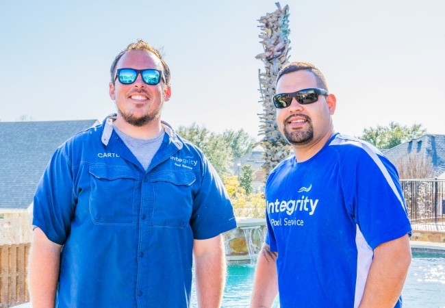 Two Integrity Pool Service employees stand smiling by a pool, wearing company shirts and sunglasses. The blue shirts are branded with the company logo. Palm tree and house in background.