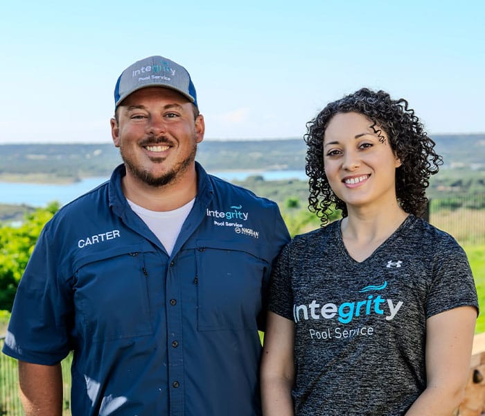 Carter and his wife, owners of Integrity Pool Service, stand outdoors. Carter wears a blue Integrity Pool Service shirt and hat. His wife wears a gray Integrity Pool Service shirt. Lake in background.