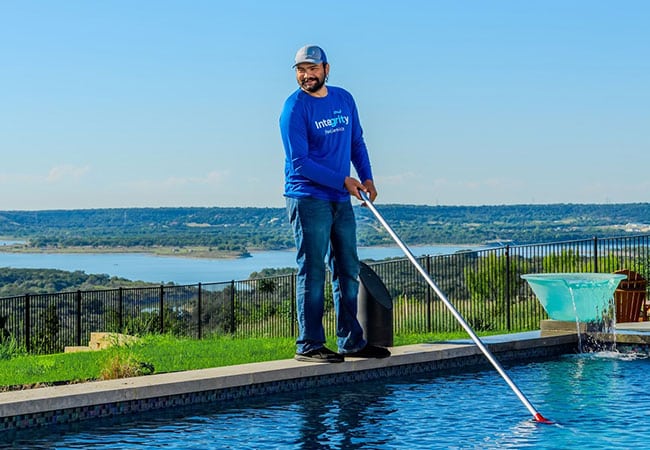 Pool service technician cleaning a pool with a long-handled brush. He wears a blue "Integrity Pools" shirt and a cap, with a scenic lake view in the background, highlighting pool maintenance careers.