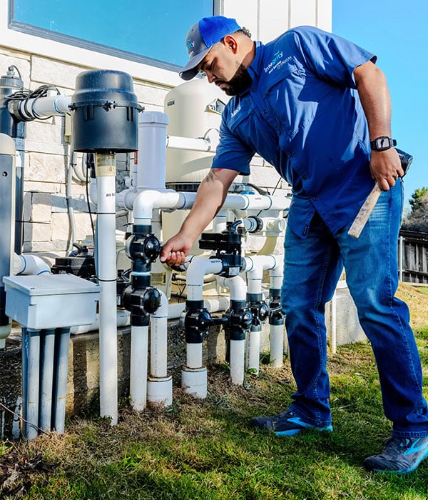 Pool maintenance technician adjusts pool filter valves. The man, wearing an "Integrity" shirt and blue cap, kneels by the pool equipment. Winter pool options include filter maintenance.