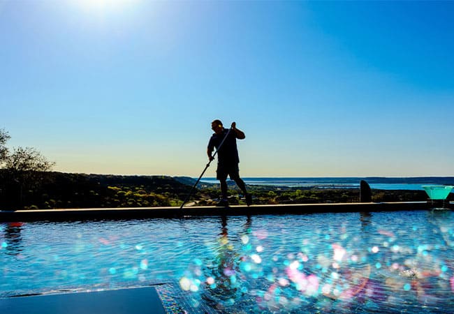 Silhouette of a person cleaning an infinity pool against a bright blue sky and scenic landscape. Sparkling water reflects the sunlight.