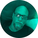 Bald man with glasses and a goatee, lying back against a dark green surface. Relaxed pose, looking at the camera.