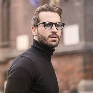 Man in black turtleneck and glasses looks over his shoulder with a concerned expression. A blurred brick building is in the background.