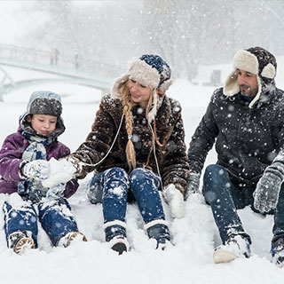 Family enjoying a snowy day: A mother, father, and child bundled in winter hats and coats, sitting in the snow. The child makes a snowball, with a bridge visible in the snowy background.