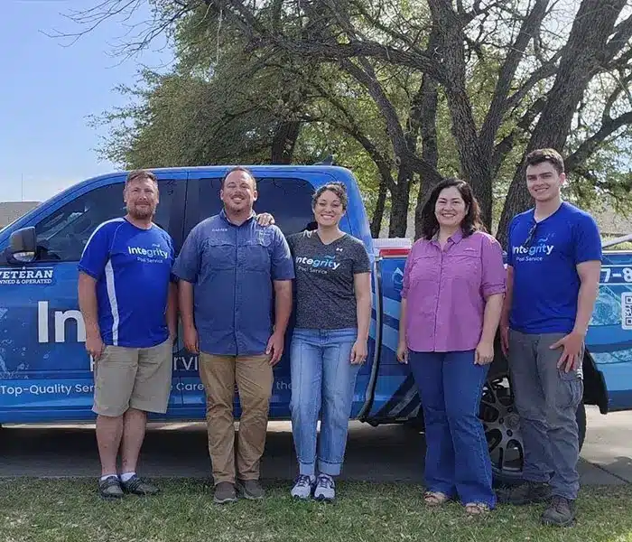 Five people standing in front of a blue branded pickup truck outdoors, smiling, wearing casual and company shirts, trees behind.