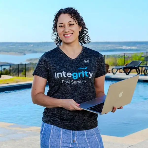 Smiling person standing beside a pool holding a laptop, wearing a dark 'Integrity Pool Service' shirt.