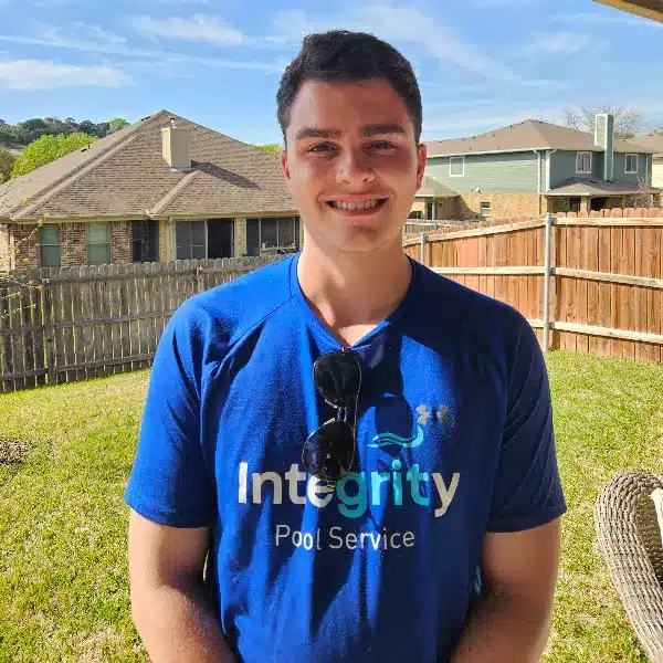 Person smiling in a sunny backyard wearing a blue Integrity Pool Service shirt with sunglasses on the collar; wooden fence and houses behind.