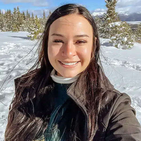 Person smiling for selfie in snowy landscape, long dark hair, dark jacket and white neck warmer, evergreen trees and sunlit mountains behind.