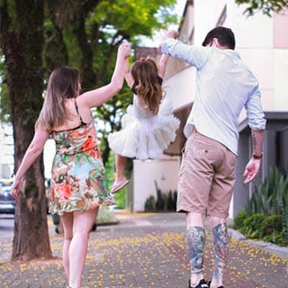 Two adults, one in a floral dress and one with tattooed legs, swing a child in a white tutu between them on a tree-lined sidewalk.