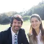 Two smiling people in business attire sitting on grass outdoors, trees and sunlight in the background.
