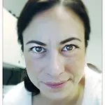 Close-up of a person with dark hair pulled back, arched eyebrows and slight smile, wearing a white top, office cabinets in blurred background