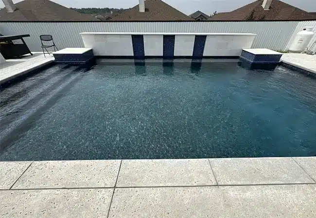 Rectangular backyard pool with dark blue tile accents, concrete deck, raised white tiled water feature, chair and rooftops in background.