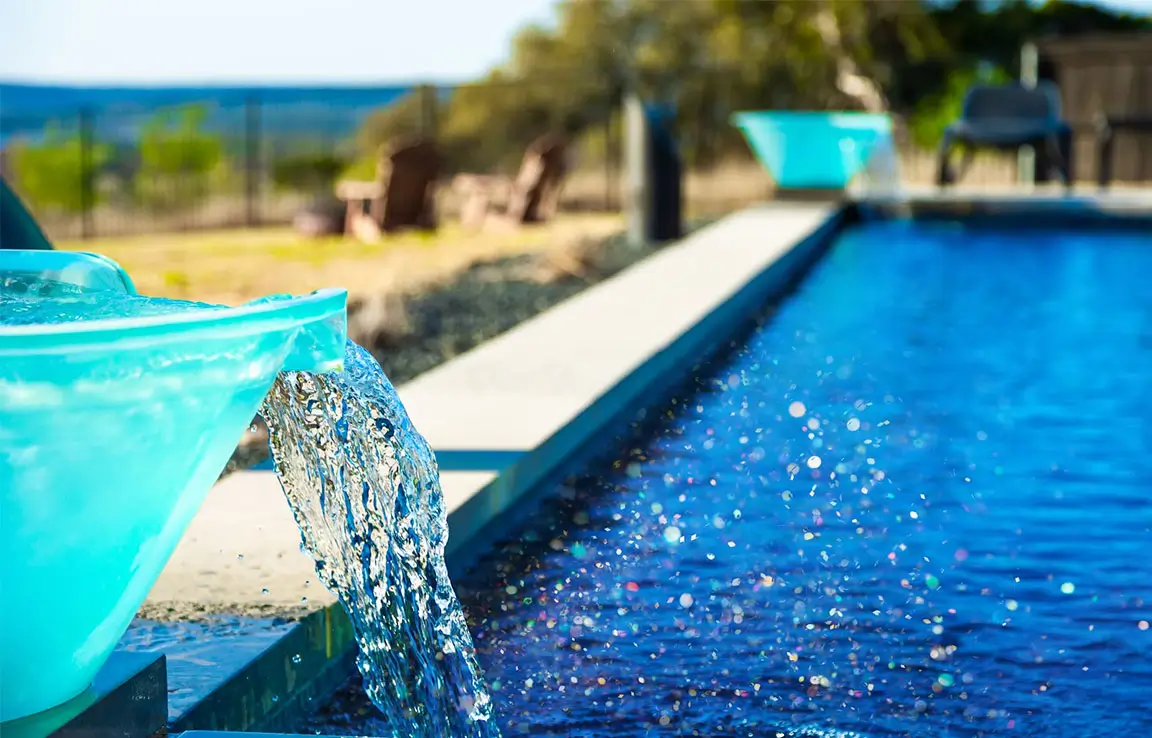 Turquoise spout pouring water into a sparkling blue pool along a concrete pool deck, blurred chairs and greenery in background.